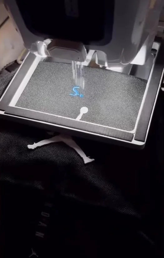 Embroidery machine adding a blue initial to black apparel.