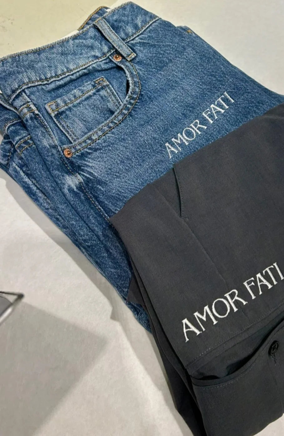 Blue jeans and dark tailored trousers embroidered with the words AMOR FATI.
