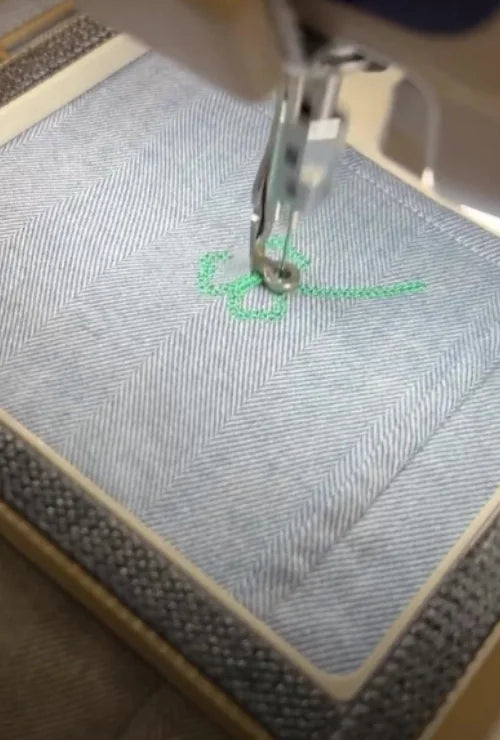 Needle stitching a small green clover motif on light herringbone fabric.