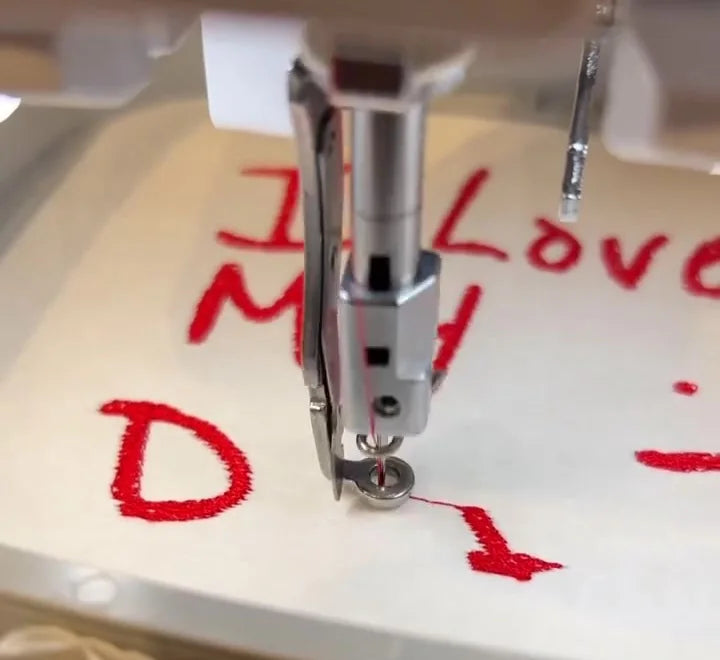 Embroidery needle stitching a red hand-drawn Father’s Day message onto white fabric.