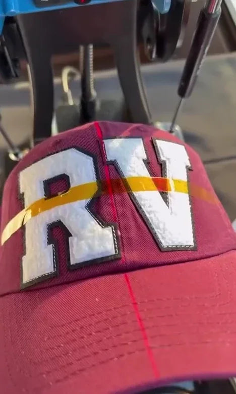 A burgundy cap with varsity-letter patches positioned at a cap press.